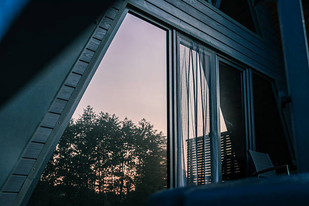 Close-up of a modern A-frame cabin window reflecting sunset and forest trees, with curtains partially drawn inside.