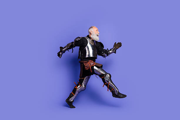 istock Elderly man dressed in shiny medieval knight armor walking confidently on a solid purple background 2221618555
