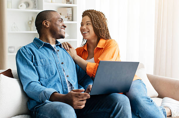 istock Black couple smiling at each other while sitting on couch with laptop 2221618562