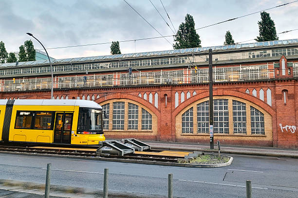 Berlin, Germany, May 25, 2025: Yellow tram at a berlin stop against a historic red brick building. overhead wires and street lamps highlight urban infrastructure. lush trees peak from behind, adding greenery to this bustling urban scene.