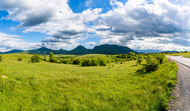 expansive meadow with low green vegetation stretching toward a mountainous backdrop. A narrow paved road runs along the right side, leading into the distance. The sky is a mix of blue and white with sunlight breaking through.