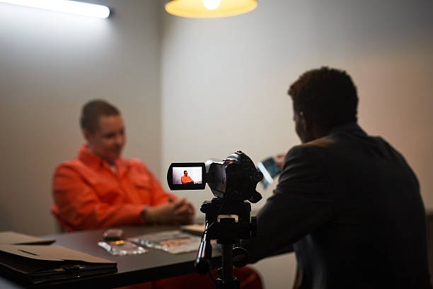 Man with a camera equipped with a microphone is conducting an interview with another man seated at a picnic table within a park gazebo, capturing video footage