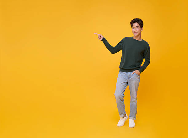 istock portrait smiling young handsome Asian man pointing finger in empty space aside isolated on yellow copy space background. 2221636170