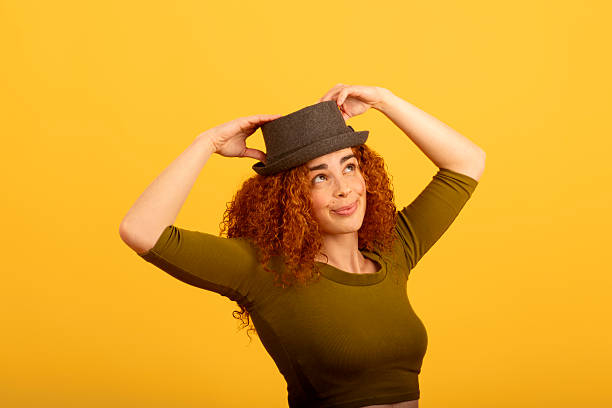 Studio shot of a cheerful redhead model adjusting her hat while smiling and looking away on a vibrant yellow background