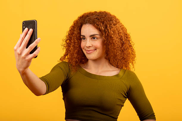 Young woman with curly red hair smiling and taking a selfie with smartphone on yellow background