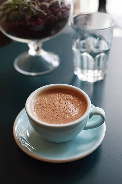 A cup of hot chocolate served in a pastel blue ceramic cup and saucer, placed on a dark teal table next to a glass of water and a decorative terrarium. Cozy and inviting atmosphere, ideal for concepts of comfort, cafe culture, and slow living.