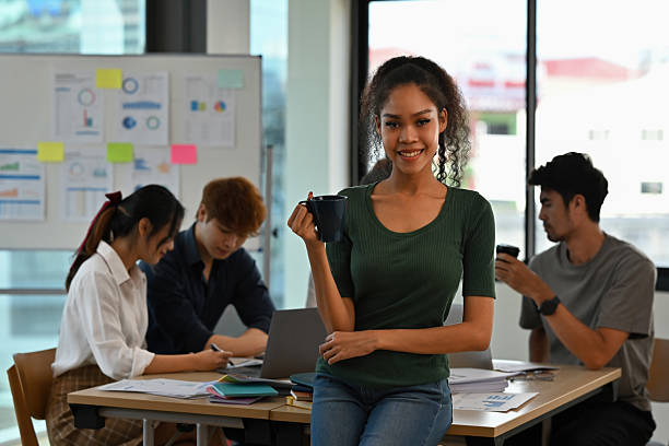 istock Confident young African woman holding a coffee mug while standing in a modern office, team members working in the background 2221645260