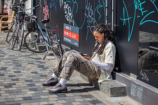 Strøget, Pedestrian Zone, Copenhagen, Zealand, Denmark - June19th 2025:  A young woman enjoys the summer sun as she sits on a doorstep outside a closed shop that's up for rent, reading a text message on her phone.