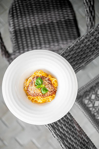 Plate of spaghetti with a grilled meat patty and fresh basil garnish served on a wicker table outdoors, evoking a cozy, rustic dining atmosphere with natural textures and soft light.