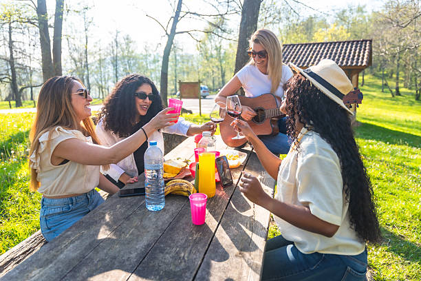 Group of female friends enjoying a picnic in the park, toasting with drinks and playing guitar