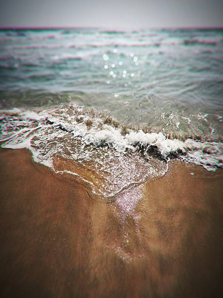 This image captures the powerful and dynamic motion of a crashing wave on the beach. The wave is breaking against the shore, creating a dramatic spray of water and foam. The image has a vibrant, almost surreal quality with the blurred, iridescent colors of the water and the textured sand in the foreground. This visually striking scene would be perfect for stock photography related to nature, the ocean, or beach themes.