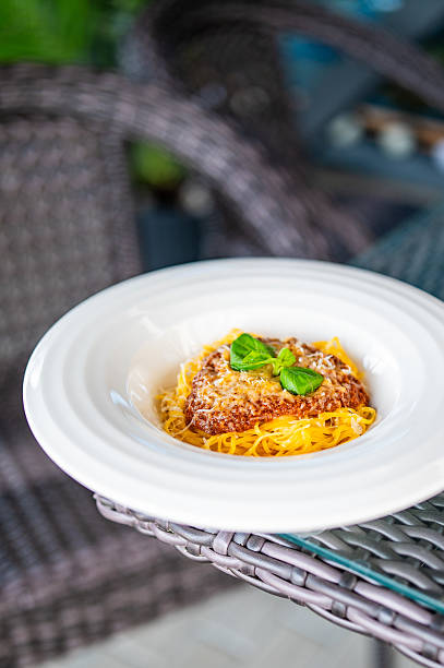 Close-up of spaghetti with meat sauce, grated cheese, and basil on a white plate atop a wicker table, outdoor setting with soft background blur, capturing a cozy Italian dining vibe.
