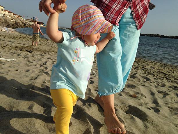 Toddler confidently grasps an adult's hand while taking first steps on a sandy beach. Another child plays near the water, creating a serene beach scene with a sense of exploration
