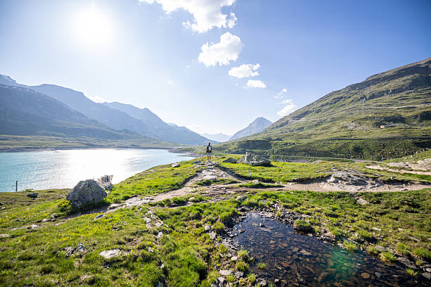Person stands on rocky path overlooking serene lake surrounded by mountains. Hiker embraces nature solitude on sunny day. Adventure and exploration in tranquil wilderness setting.