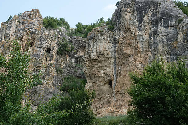A breathtaking landscape featuring towering rock formations with prominent cliffs and natural caves. Lush green vegetation thrives at the base and along the cliffs, contrasting the rugged stone.