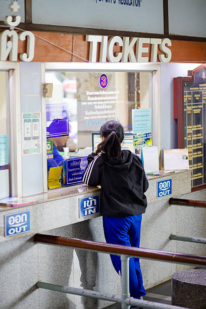 Young thai woman is buying a train ticket at counter desk in station of Lampang. Lampang is a city in north Thailand south of Chiang Mai. Woman is wearing casual clothes and has long natural black hair. She is communicating with staff inside the counter office behind the glass. Man working with pc and is showing train schedules and tickets on screen to the woman. Rear view of standing woman at counter