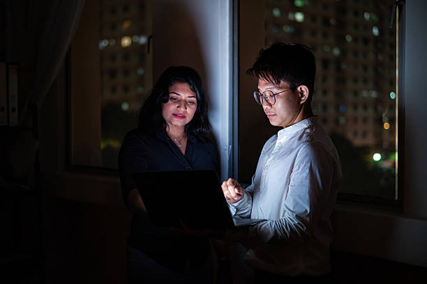 istock Two Asian business colleagues having conversation and discussing office work using a laptop at the workplace during night hours. 2221720354