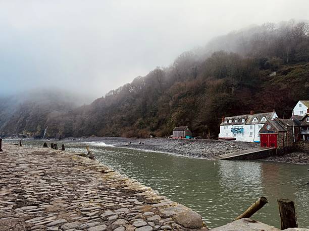 A quiet, atmospheric view of Clovelly Harbour on a misty morning. The cobbled slipway leads down to calm harbour waters, while traditional white buildings cling to the hillside in the background. This historic fishing village in North Devon is known for its steep cobbled streets, timeless charm, and vehicle-free tranquility.