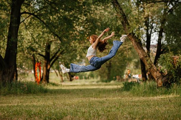 A woman in casual attire performs an impressive leap mid-air in a lush park. Her expression reflects joy and freedom, capturing the essence of movement and nature.