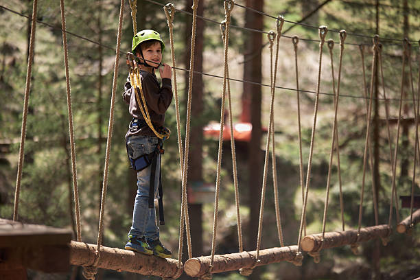 Boy wearing a green helmet and safety gear looks into the camera while standing on a log in a forest playground.