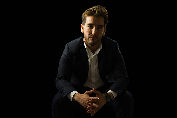 Confident businessman sitting with hands clasped, wearing suit and white shirt on dark background