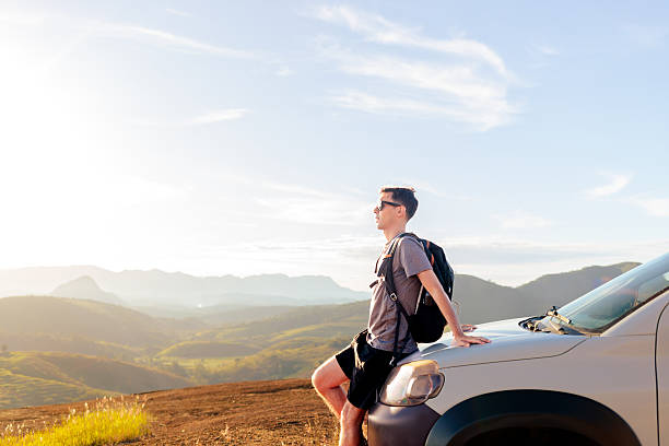 Traveler leaning on his car at sunset, looking at the mountains. A moment of freedom, inspiration, and connection with nature during a road trip.