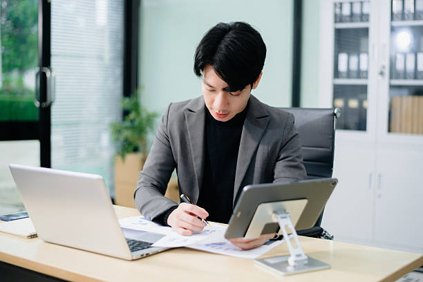 istock Young Asian businessman in suit smiling and talking on phone while working on charts with laptop and tablet. 2221784343