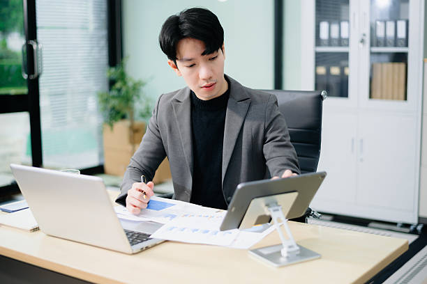 istock Young Asian businessman in suit smiling and talking on phone while working on charts with laptop and tablet. 2221784349