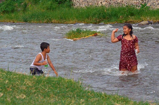A boy and girl walk and play in the river stream holding plastic bottles, having fun together in the cool flowing water, surrounded by nature.