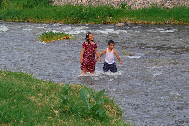 Two children stand in the river, gazing calmly at the current, surrounded by lush grass and wild plants lining the riverside.