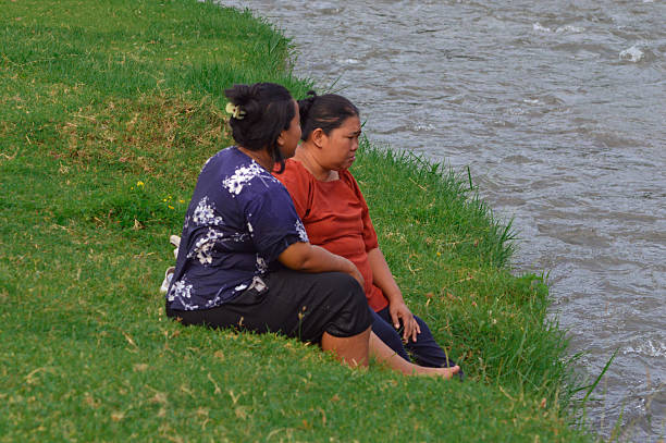 Two women sit and chat on a grassy slope beside the stream, appearing peaceful and enjoying a quiet moment near the gently flowing water.