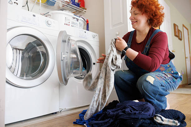 Mid-adult redhead woman smiles while removing laundry from dryer in home utility room. 47-year-old woman wearing denim overalls and burgundy shirt kneels beside front-loading washer and dryer set. She holds gray fabric while pile of dark clothes sits on wooden floor. Smart watch visible on wrist. Utility shelving with household supplies visible above appliances.