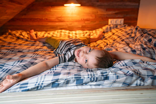 A cheerful boy lies on a bed with arms outstretched, enjoying warmth and relaxation. The setting is cozy with soft light illuminating the wooden textures and plaid bed linens.