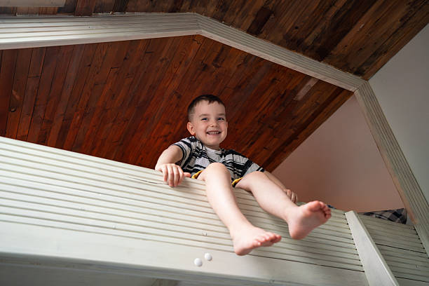 Cheerful young boy enjoys sitting playfully on a bright bunk bed, smiling warmly against a wooden ceiling backdrop, radiating joyful innocence and carefree delight.