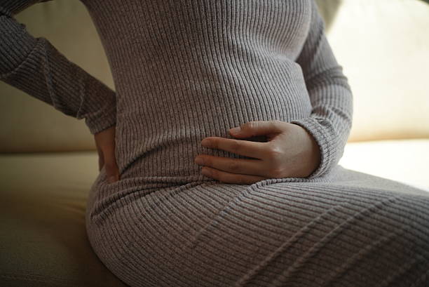 istock Close up discomfort pregnant woman touching lower back by suffering backache while sitting uncomfortably on sofa at home, prenatal care and health check up concept. 2221804762