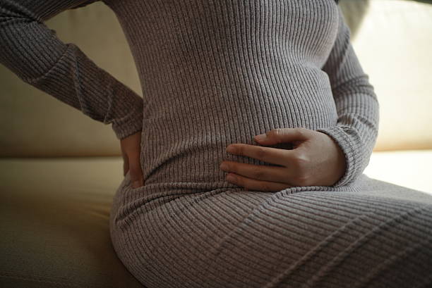 istock Close up discomfort pregnant woman touching lower back by suffering backache while sitting uncomfortably on sofa at home, prenatal care and health check up concept. 2221804772