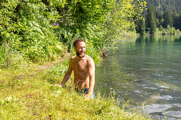 Man with beard enjoying a swim in clear lake surrounded by lush greenery. Nature enthusiast exploring serene outdoor settings. Relaxing moment in untouched wilderness by the water.