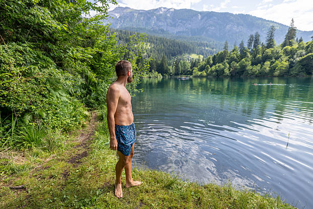 Shirtless man stands by the tranquil lake, surrounded by lush forest and mountains. Peaceful scene with nature's beauty. Ideal for outdoor adventure or nature photography.