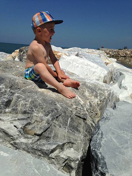 Child sitting on large coastal rocks, smiling and enjoying a sunny day by the sea. Wearing a striped cap and shorts, the child exhibits joy and curiosity amidst the natural landscape.