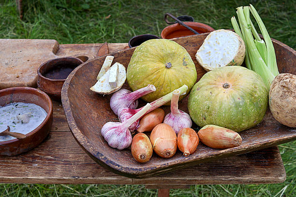 Rustic wooden bowl filled with vegetables including garlic, onions, squash, and celeriac arranged on a wooden table outdoors. A bowl of creamy soup and rustic kitchen tools add to the rural setting.