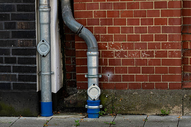 A metallic water pipe system is installed next to a red brick wall. The gray and blue pipes are connected, showcasing urban infrastructure in a city setting. Greenery peeks through the pavement.
