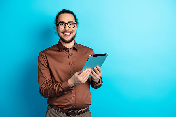 istock Young businessman smiling confidently while holding a tablet against a teal background, showcasing modern professional style 2221835542