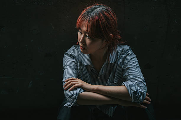 Portrait of a non-binary Japanese person seated indoors in low-key lighting, glancing to the side with a contemplative expression. The contrast between shadows and highlights enhances the cinematic tone, evoking a sense of inner thought, isolation, or personal reflection. The subject wears a striped shirt and sits against a dark concrete wall, capturing themes of emotional depth, identity, and quiet resilience.