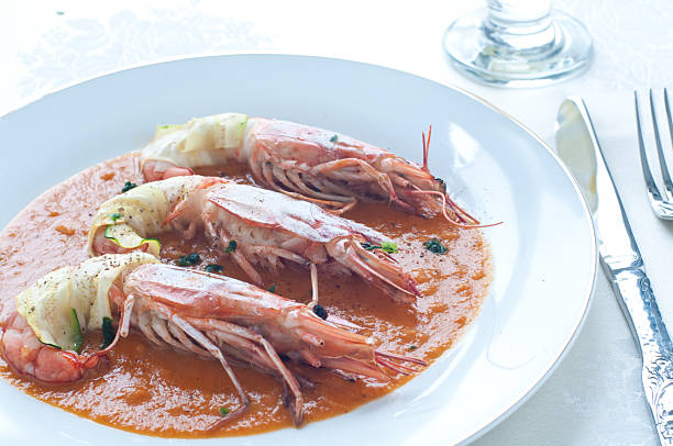 The soup is presented in a white ceramic bowl, filled with a rich, orange-red broth that contains various fresh seafood ingredients, such as shrimp and scallops.The accompanying bread slice has been toasted to a golden-brown hue and appears to be generously topped with a savory garlic butter or compound butter. This warm, aromatic bread is perfectly suited for dipping into the bold and hearty seafood tomato soup.
