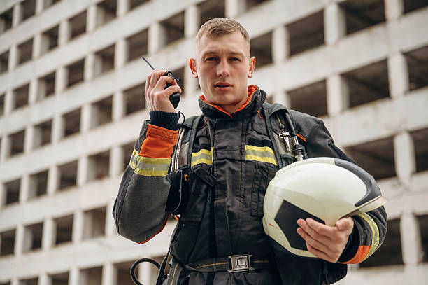 istock Conversation by walkie talkie. Professional firefighter is standing outdoors against big unfinished building 2221874299
