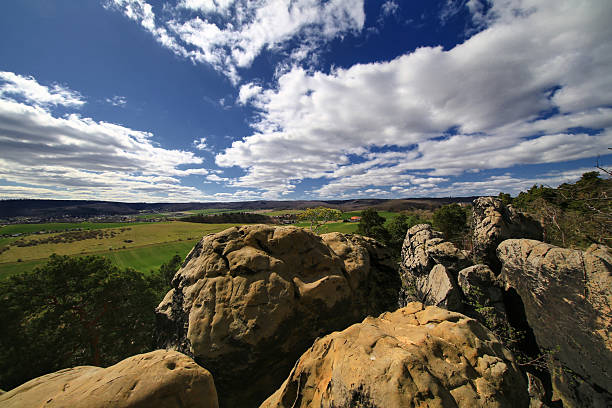 Sandstone cliff edge in the Harz mountains overlooking green fields and hills under a wide blue sky with broken clouds.