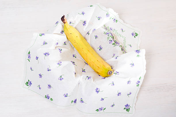 A single ripe yellow banana rests on a white floral patterned cloth napkin on a light wooden table. The napkin is folded around the fruit.