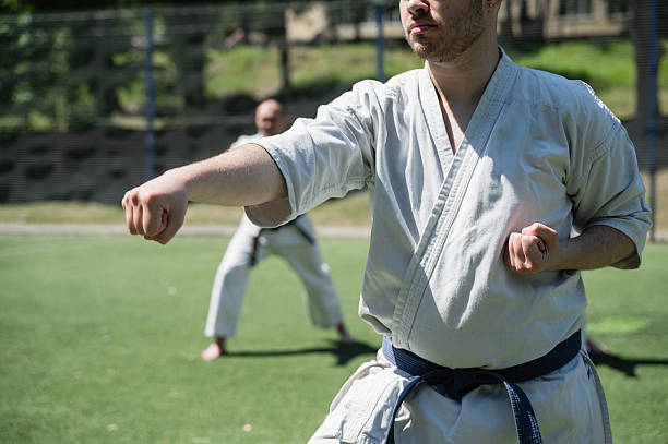 Man in white karate gi and blue belt performing martial arts technique on outdoor field, another person blurred in background, sunny weather, close-up.
