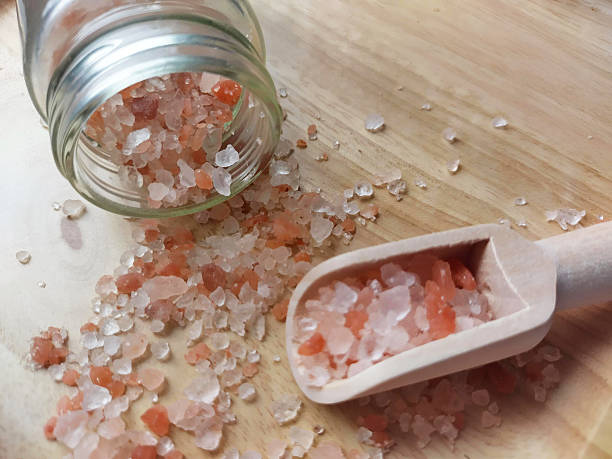 Pink Himalayan Salt Crystals Spilled from a Jar, with a Wooden Scoop on a Textured Surface.