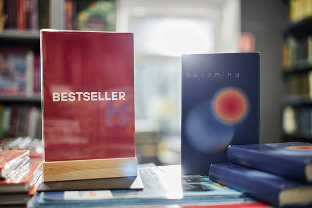 Stack of books on display in bookstore with red book labeled Bestseller standing upright next to blue book titled Becoming, blurred shelves filled with books in background
